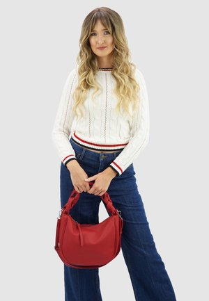 Rode leren handtas met een gevlochten handvat, met een gladde textuur en afgeronde vorm. Model draagt een witte kabelgebreide trui en blauwe spijkerbroek.