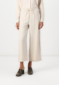 Celana lebar berwarna beige dengan pinggang tali, terbuat dari bahan tekstur. Dipadukan dengan sepatu mules bertumit blok berlubang berwarna gelap.