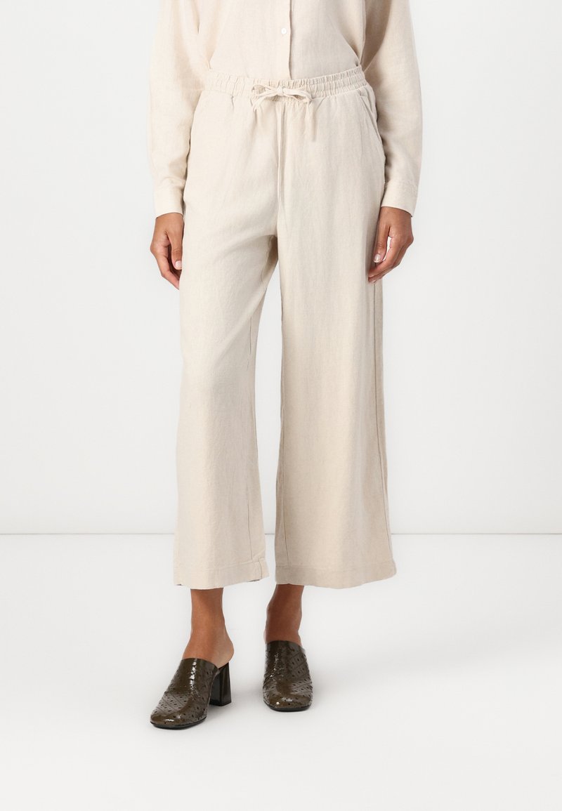 Celana lebar berwarna beige dengan pinggang tali, terbuat dari bahan tekstur. Dipadukan dengan sepatu mules bertumit blok berlubang berwarna gelap.