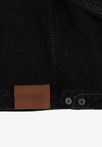 Czarna tkanina dżinsowa ma skórzaną metkę z wytłoczonym napisem "PROSTO". Dwa srebrne guziki są widoczne w pobliżu dołu, kontrastując z ciemnym materiałem.