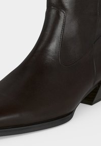 Gros plan d’une botte cheville en cuir marron foncé avec un bout pointu et un petit talon empilé sur un fond clair.