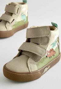 Scarpe alte color tan con doppie cinghie in Velcro, decorate con stampe di dinosauri e suola in gomma testurizzata. Fodera in tessuto morbido e linguetta per un facile indossamento.
