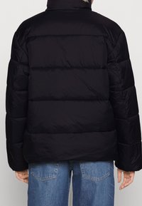 Svart puffjacka med hög krage, med kviltade sektioner, slät yta och något överdimensionerad passform. Tillsammans med blå denim.