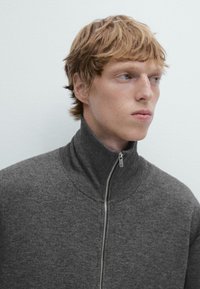 Maglione grigio con zip e collo alto, realizzato in un tessuto morbido e texture. Presenta una zip metallica e una silhouette aderente.