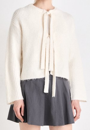 Vrouw draagt een witte geribbelde gebreide trui met strikjes aan de voorkant en een geplooide donkergrijze mini-rok tegen een effen achtergrond.