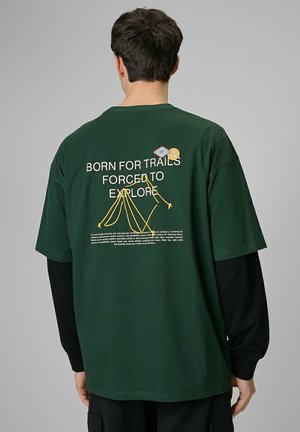 Mężczyzna noszący zieloną koszulkę z krótkim rękawem na czarnych długich rękawach z żółtym haftem namiotu i napisem „Born for trails, forced to explore” na plecach.