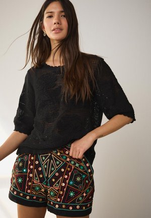 Femme portant un haut noir texturé et un short coloré brodé, debout sur un fond clair uni, avec de longs cheveux lâchés.