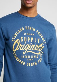 Blå sweatshirt med rund grafisk tryck och gul text: "Supply Originals" samt "Standard Denim Product, etablerad 1990."