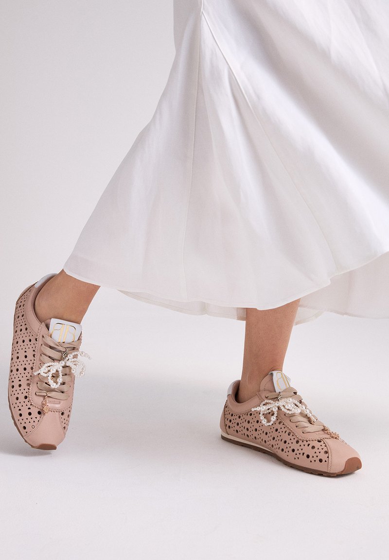 Pies con zapatillas beige perforadas y cordones decorados con perlas, caminando con una falda blanca fluida en un entorno de estudio.