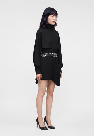 Femme aux longs cheveux foncés porte une mini-robe noire à manches longues avec un col roulé, une ceinture à clous argentés et des talons hauts noirs, debout sur fond blanc.