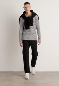 Svart-hvitt stripet langermet skjorte, rund hals, med oransje detaljer, kombinert med svarte bukser og hvite sneakers. Glatte teksturer er synlige.