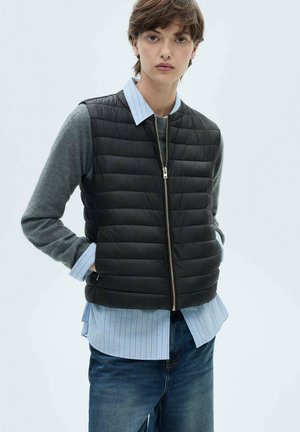 Zwarte gequiltte gilet met ritssluiting, gedragen over een grijs shirt met lange mouwen en een blauw gestreept overhemd, in combinatie met een blauwe spijkerbroek.