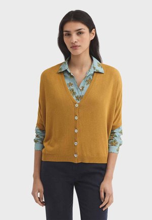 Vrouw draagt een mosterdgele gebreide cardigan met knopen over een blauw bloemenblouse en donkere broek, staand tegen een effen achtergrond.