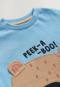 Svetlomodrá košeľa s krátkym rukávom s dizajnom medvedej tváre v hnedej farbe, vyšívanými detailmi a nápisom "PEEK-A-BOO!" na prednej strane.