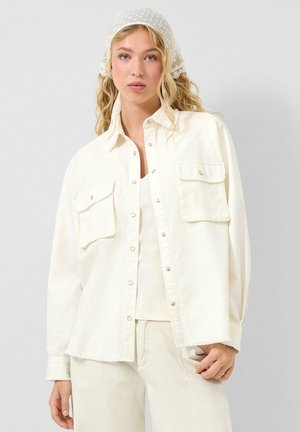 Jonge vrouw met golvend blond haar, gekleed in een crème oversized blouse met knopen, bijpassende broek en een licht polkadot hoofddoekje.