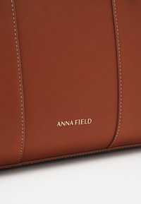 Sac en cuir marron avec un marquage doré "ANNA FIELD", orné de coutures claires et d'une texture lisse et raffinée.
