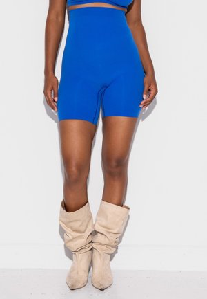 Kvinde iført lyseblå højtsiddende biker-shorts og beige slaskede støvler til midt på læggen, stående op ad en hvid væg.