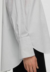 Camicia bianca con sottili righe verticali nere, dotata di collo a punta e maniche ampie raccolte ai polsini.