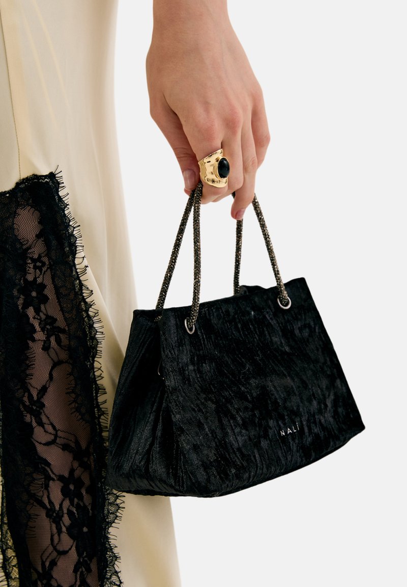 Czarna welurowa torebka o fakturowanej powierzchni, plisowanym kształcie i krótkich, metalicznych uchwytach w formie łańcuszka. Złoty pierścionek na ręce z owalnym czarnym kamieniem.