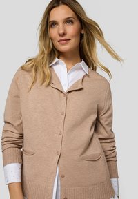 Beige Strickjacke aus weichem Strickmaterial mit durchgehender Knopfleiste, zwei Fronttaschen sowie geripptem Saum und Bündchen, über einem weißen Hemd getragen.