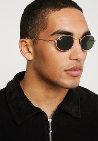 Occhiali da sole rotondi neri con montatura in metallo dorato, caratterizzati da un design elegante e lenti scure. Il modello indossa una giacca nera con colletto.
