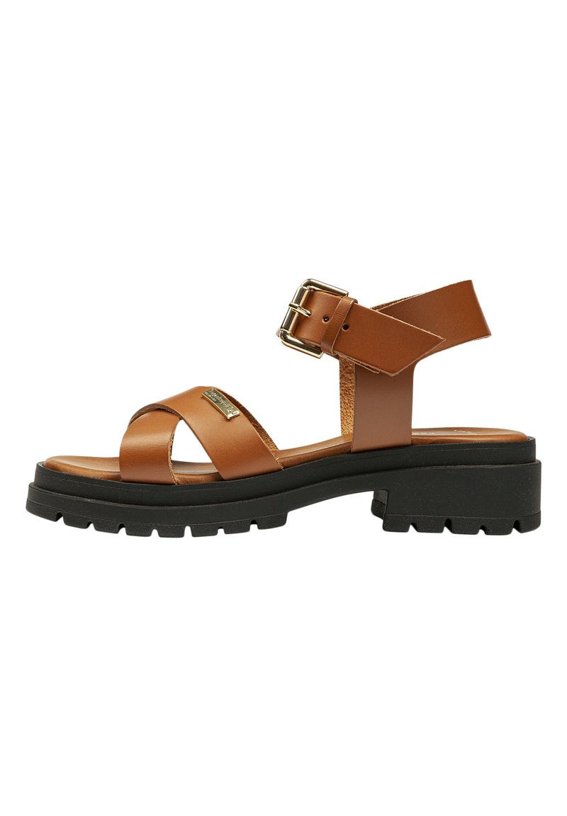 Brun lædersandal med to krydsende stropper, en ankelrem med en guldbuckle, og en kraftig sort gummisål.