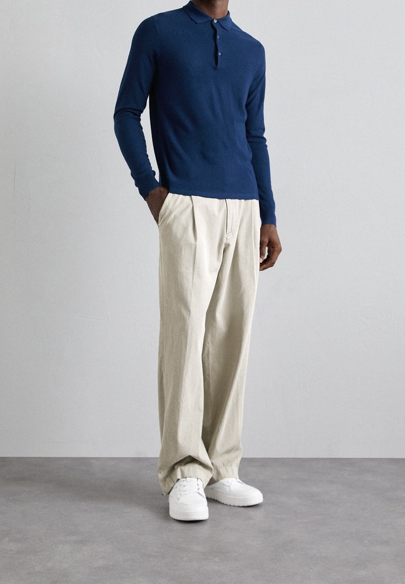 Homme portant un polo bleu marine à manches longues, un pantalon beige à jambes larges et des baskets blanches, debout devant un fond clair uni.