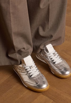 Zilveren metallic sneakers met een gladde textuur, witte veters en een gumrubberen zool, gedragen onder bruine, relaxed-fit broeken.