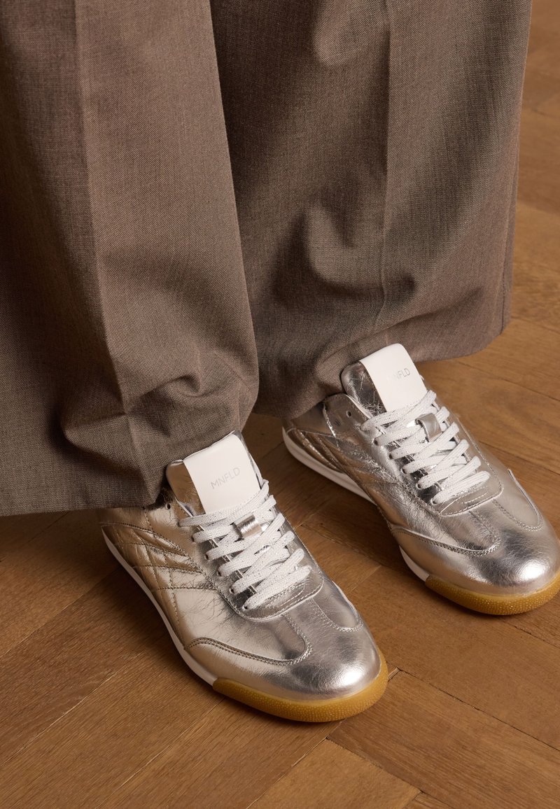 Zilveren metallic sneakers met een gladde textuur, witte veters en een gumrubberen zool, gedragen onder bruine, relaxed-fit broeken.