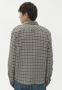 Mężczyzna w brązowej, białej i czarnej koszuli w kratę z długim rękawem oraz niebieskich dżinsach, zwrócony plecami w stronę jednolitego białego tła.