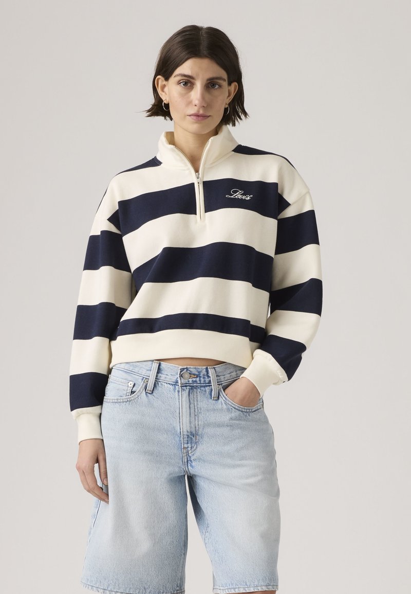 Γυναίκα με κοντά σκουρόχρωμα μαλλιά, που φοράει λευκό και ναυτικό μπλε φούτερ με φερμουάρ έως το στήθος και ανοιχτόχρωμο τζιν σορτς, στέκεται με το ένα χέρι στην τσέπη.