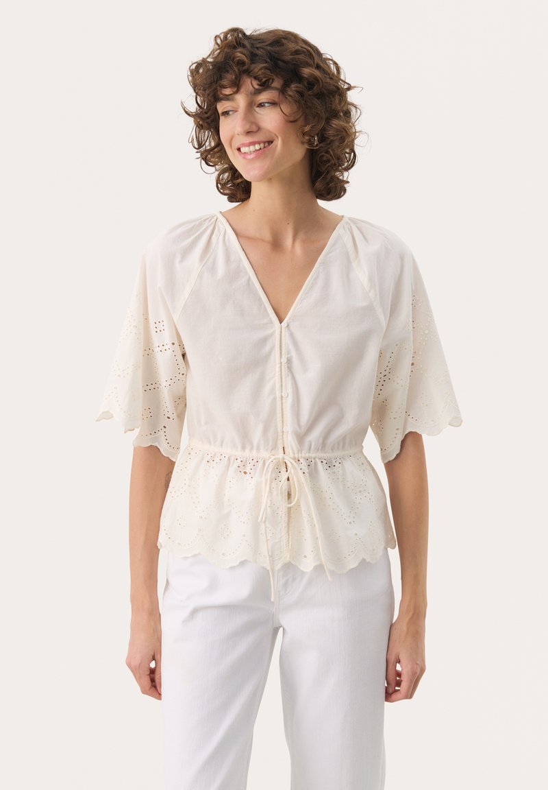Elfenbensfarvet bluse med korte ærmer, V-hals, eyelet-detaljer, snor i taljen og bølget kant, parret med hvide bukser.