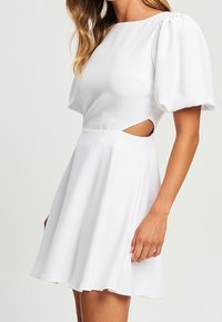 Valkoinen lyhyt mekko, jossa on puffihihat, istuva vartalo ja sivuleikkaukset. Keinotteleva hame, sileä texture ja kevyt materiaali mukavuutta varten.