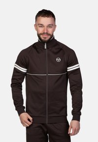 Veste zippée marron avec un col haut, rayures horizontales blanches sur les manches et un logo circulaire sur la poitrine. Tissu lisse, design ajusté.