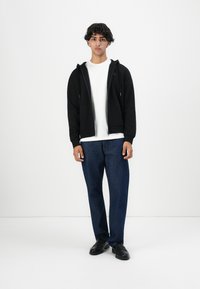 Ung mann som står i uformell antrekk med svart zip-up hettegenser, hvit t-skjorte, blå jeans og svarte sko mot en ensfarget hvit bakgrunn.