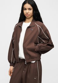 Νεαρή γυναίκα ντυμένη με καφέ φόρμα Puma με λευκές ρίγες και λευκό πουκάμισο, στέκεται με το ένα χέρι στην τσέπη του μπουφάν της.