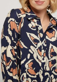 Robe bleu marine à boutons avec des motifs abstraits beiges et rouille, dotée d'un col et d'un détail de jupe plissée, fabriquée en tissu léger.