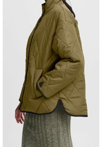 Donna che indossa una giacca trapuntata verde oliva con tasche laterali e rifiniture nere sopra una gonna verde a coste, vista di profilo.