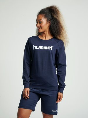 Vrouw met krullend haar die een marineblauw hummel sweatshirt en bijpassende korte broek draagt, staand en glimlachend terwijl ze opzij kijkt.