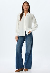 Witte blouse met een relaxed fit en geplooide mouwen, gecombineerd met een wijdvallende blauwe spijkerbroek en bruine haklaarzen. Eenvoudig en veelzijdig ontwerp.