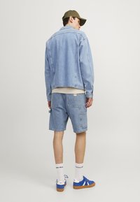 Giubbotto di jeans blu chiaro con una vestibilità rilassata, abbinato a pantaloni shorts in denim dello stesso colore. Scarpe da ginnastica blu e calzini bianchi con testo completano il look.