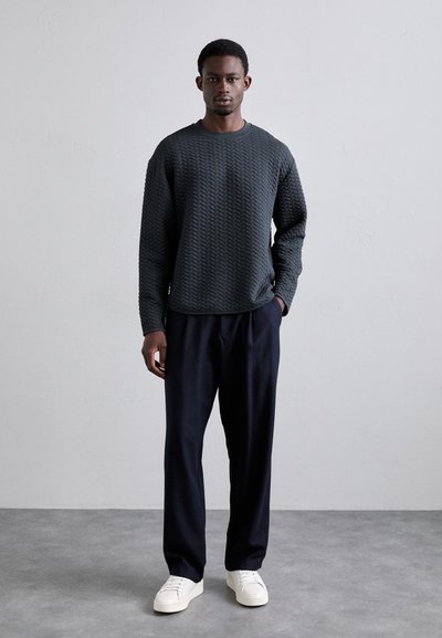 Sweatshirt texturé gris foncé avec un col rond et des manches longues, assorti à un pantalon bleu marine ample et des baskets blanches.
