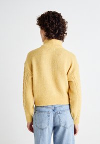 Pulover tricotat galben cu guler înalt și detalii cu model de cablu pe mâneci. Purtați cu blugi albaștri deschiși. Aspect texturat și confortabil.