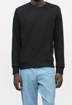 Man draagt een effen zwart sweatshirt met lange mouwen en lichtblauwe spijkerbroek, staande tegen een eenvoudige witte achtergrond.