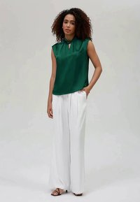 Blusa verde de cetim sem mangas com decote em gota, combinada com calças largas brancas, apresentando uma textura suave e um design minimalista.