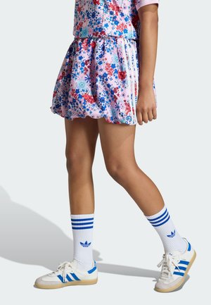 Vestito rosa con motivo floreale, maniche corte, gonna arricciata e tre strisce bianche. Abbinato a calzini a righe blu e bianche e scarpe da ginnastica.