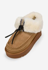 Zapato de casa estilo mocasín de color marrón, con suela de goma gruesa, cuello de borreguito cremoso y esponjoso, ribete decorativo cosido y lazada negra.