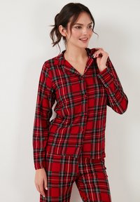 Punainen ruudullinen pyjama-asuste sisältää nappipaidan, jossa on kaulus ja pitkät hihat, sekä siihen sopivat housut, jotka on valmistettu pehmeästä kankaasta.