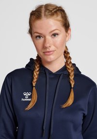 Felpa blu navy con tasca a marsupio frontale, texture leggermente lucida, cappuccio con cordoncino e logo "hummel" bianco sul petto.