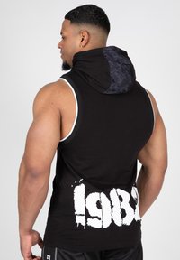 Svart ärmlös hoodie med ett mönstrat grått huva, som har en stor vit "1982" grafik på baksidan och vita detaljer längs armhålen.